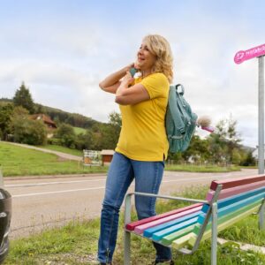 Eine blonde Frau mit Rucksack steht vor einer bunten Bank und lächelt zu einem schwarzen Auto, das nur wenig von vorne zu sehen ist. Rechts von der Bank steht ein Schild mit der Aufschrift: Mitfahrbänkle.